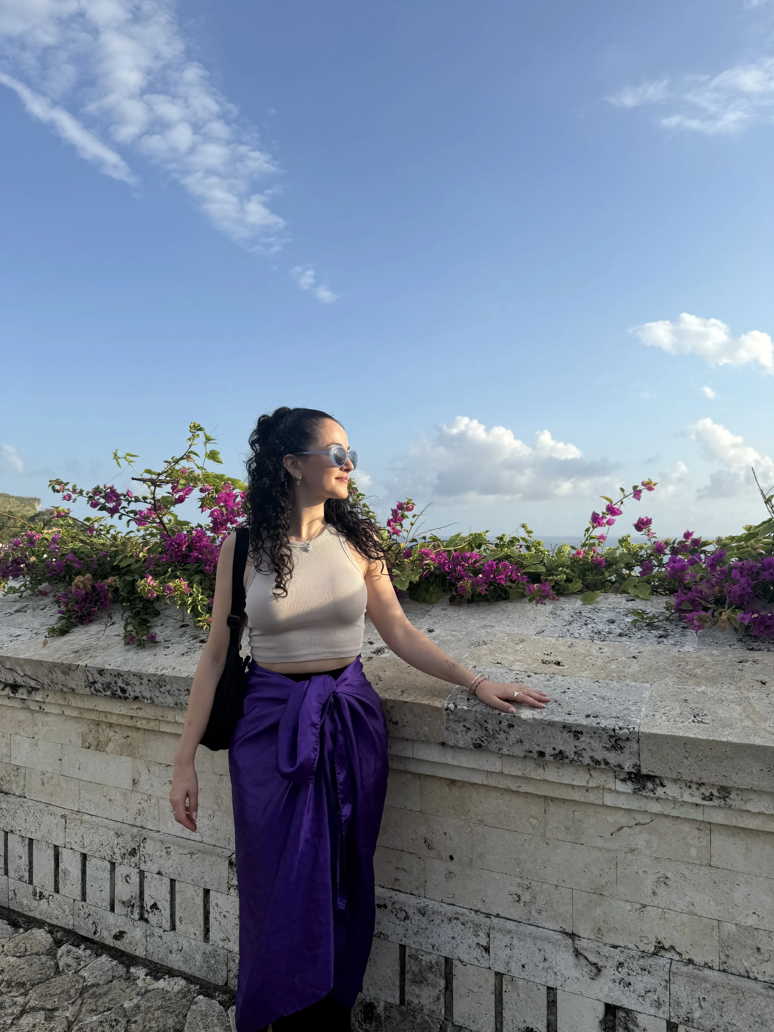 A woman with curly black hair and sunglasses leaning on a stone wall with pink flowers, under a blue sky with scattered clouds.