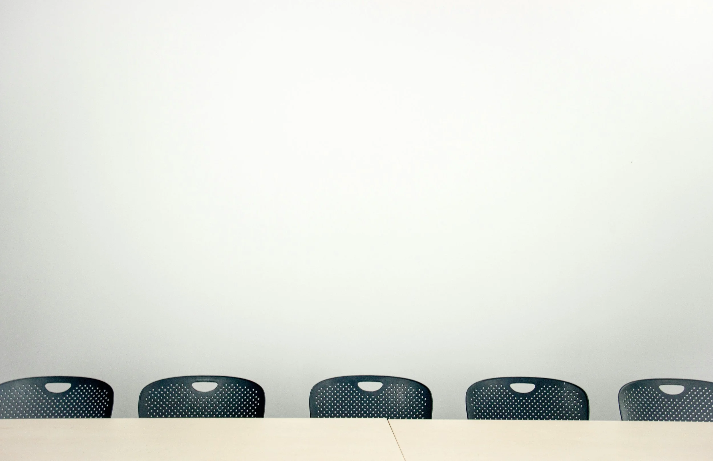 Four black chairs with perforated backs lined up against a plain white wall and a long white table in front of them.