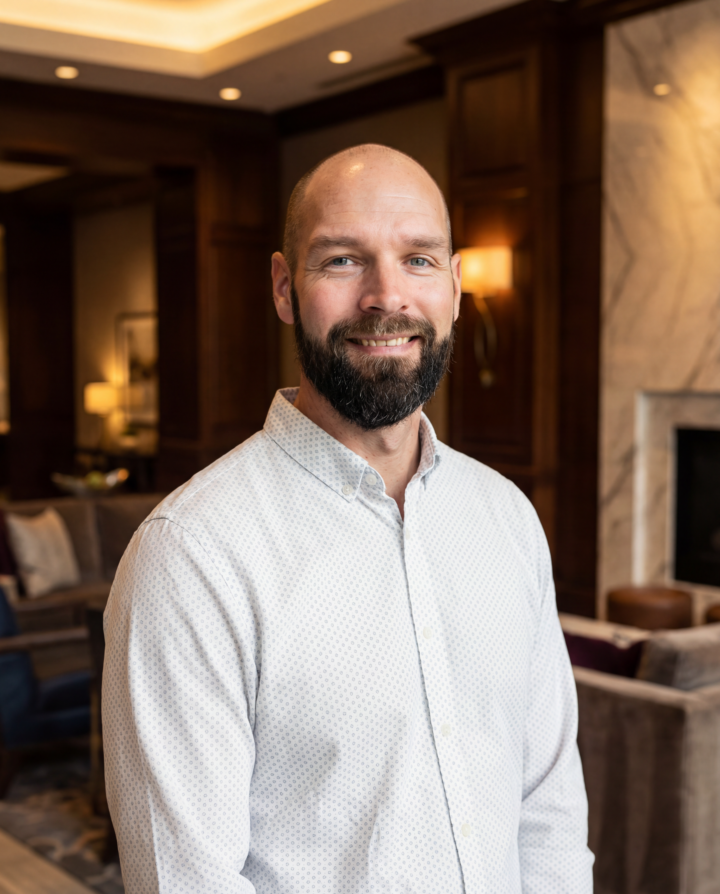 A smiling man with a beard in a white button-up shirt standing in a warmly lit room with wooden paneling and a fireplace.