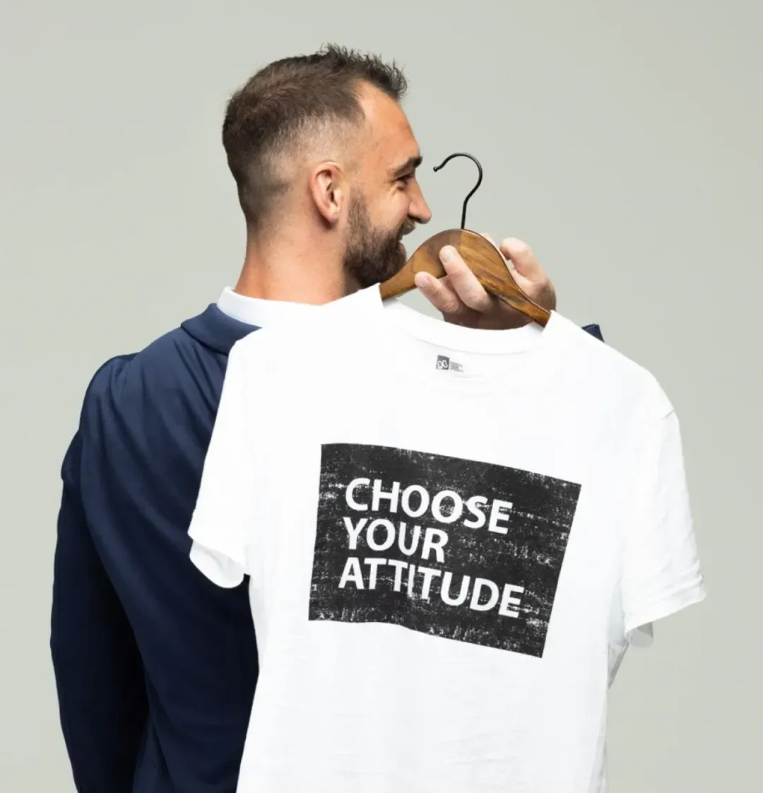 A man with a beard holding a white t-shirt that reads 'Choose Your Attitude' on a hanger, smiling and appearing to smell or enjoy the shirt.