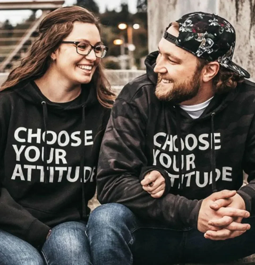 A young woman and man sitting together outdoors, smiling and looking at each other, both wearing black hoodies with the phrase 'Choose Your Attitude' printed on them.