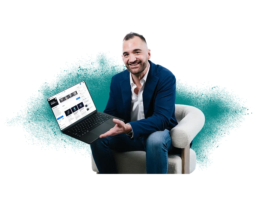 A man in a blue blazer and white shirt sitting on a white armchair, holding a laptop that displays a website, smiling at the camera.