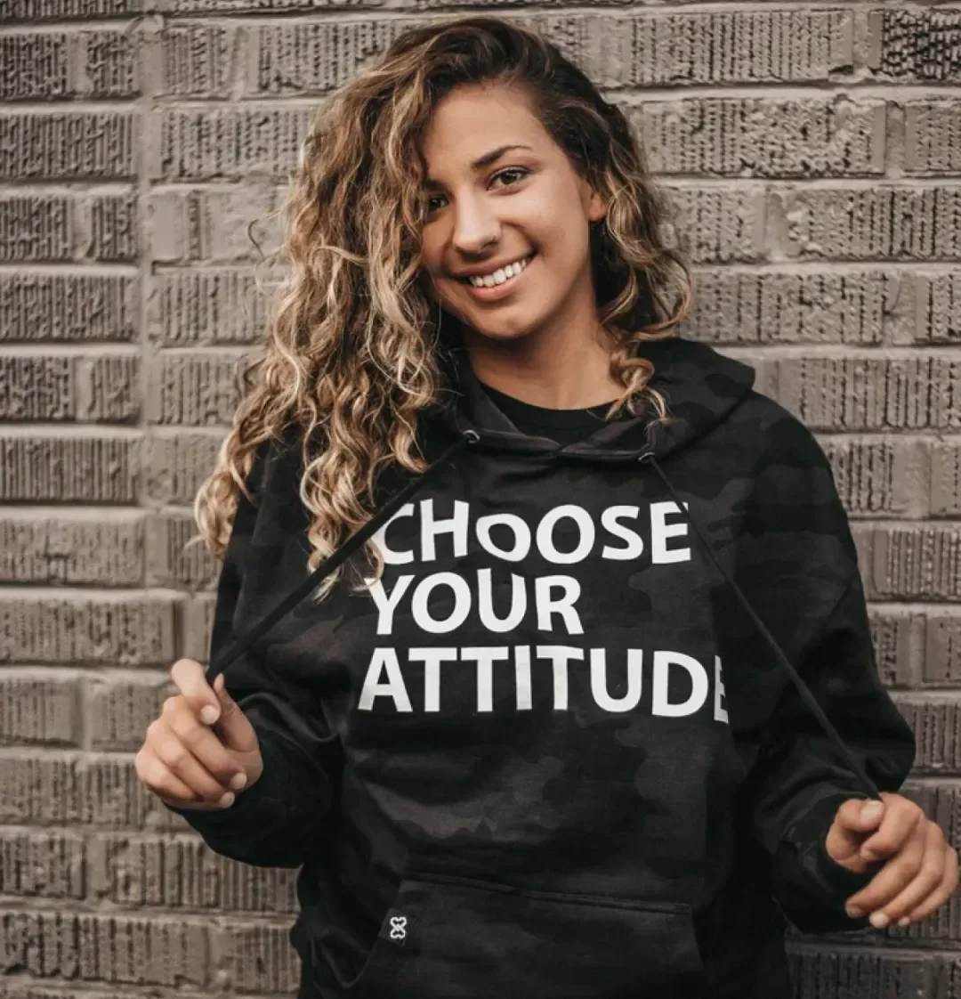 A young woman with curly hair smiling and holding the strings of her black hoodie, which has the message "CHOOSE YOUR ATTITUDE" printed on it, standing against a brick wall.