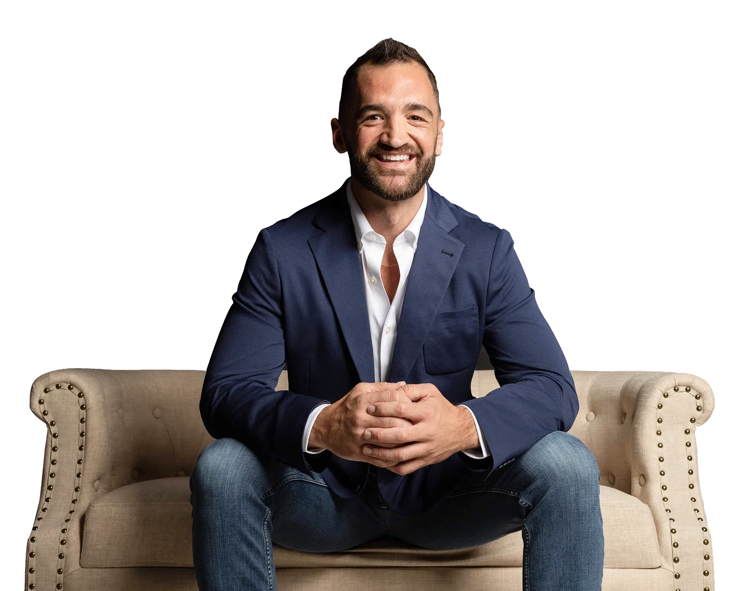 A smiling man with a beard sitting on a beige sofa, wearing a navy blazer and jeans, with his hands clasped in front of him.