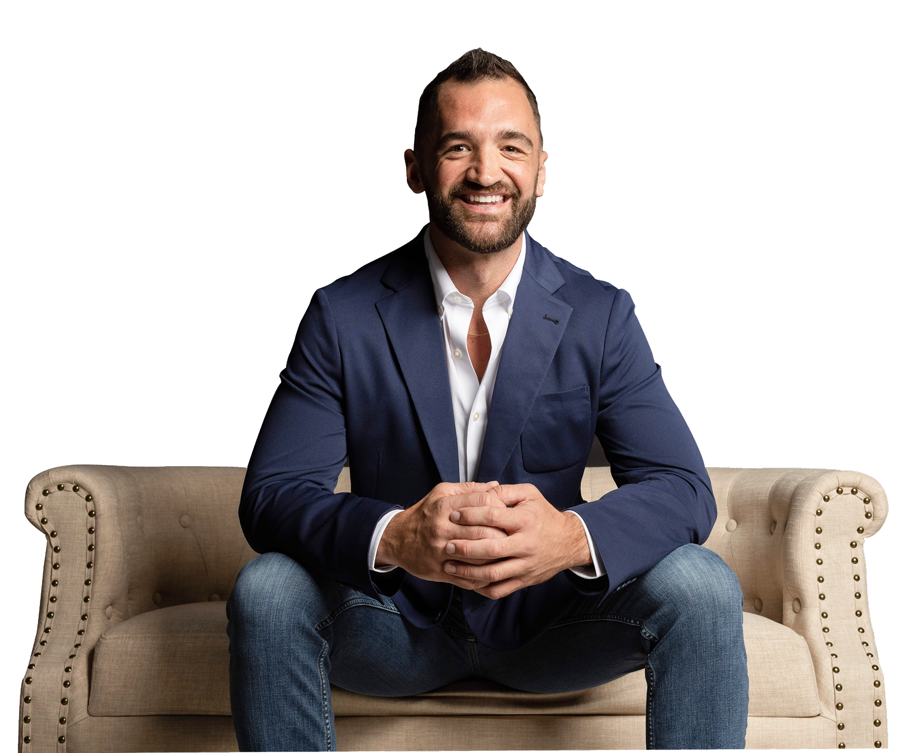 A smiling man in a blue blazer and white shirt sitting on a beige tufted sofa with brass nailhead trim.
