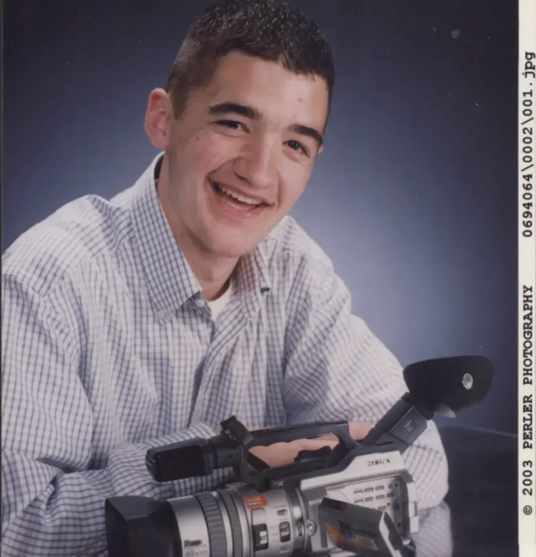 Young man with short dark hair smiling, holding a professional video camera, wearing a checkered shirt against a dark backdrop.