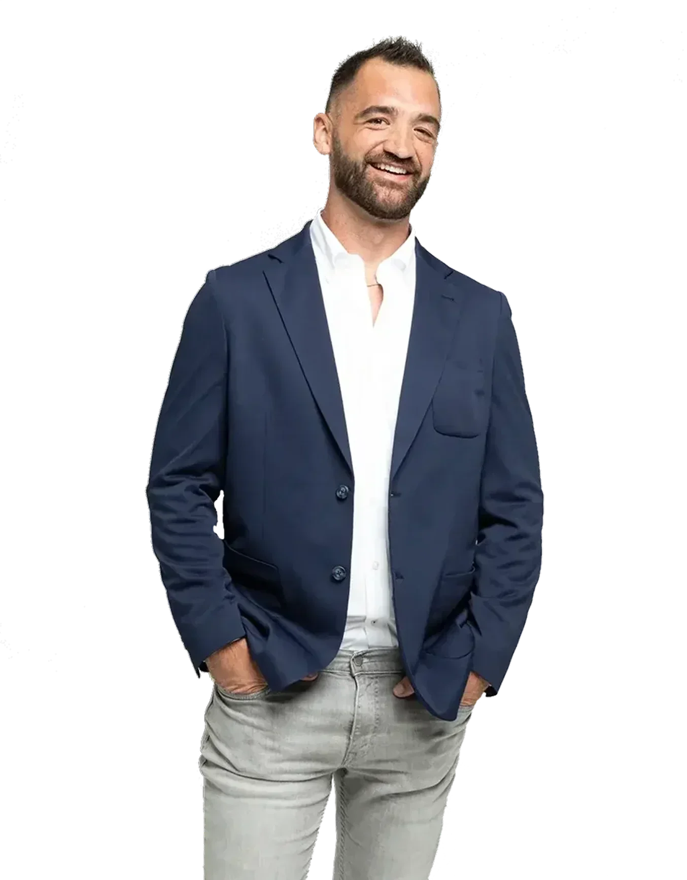 A man smiling, wearing a navy blue blazer, white shirt, and light-colored pants with hands in pockets.