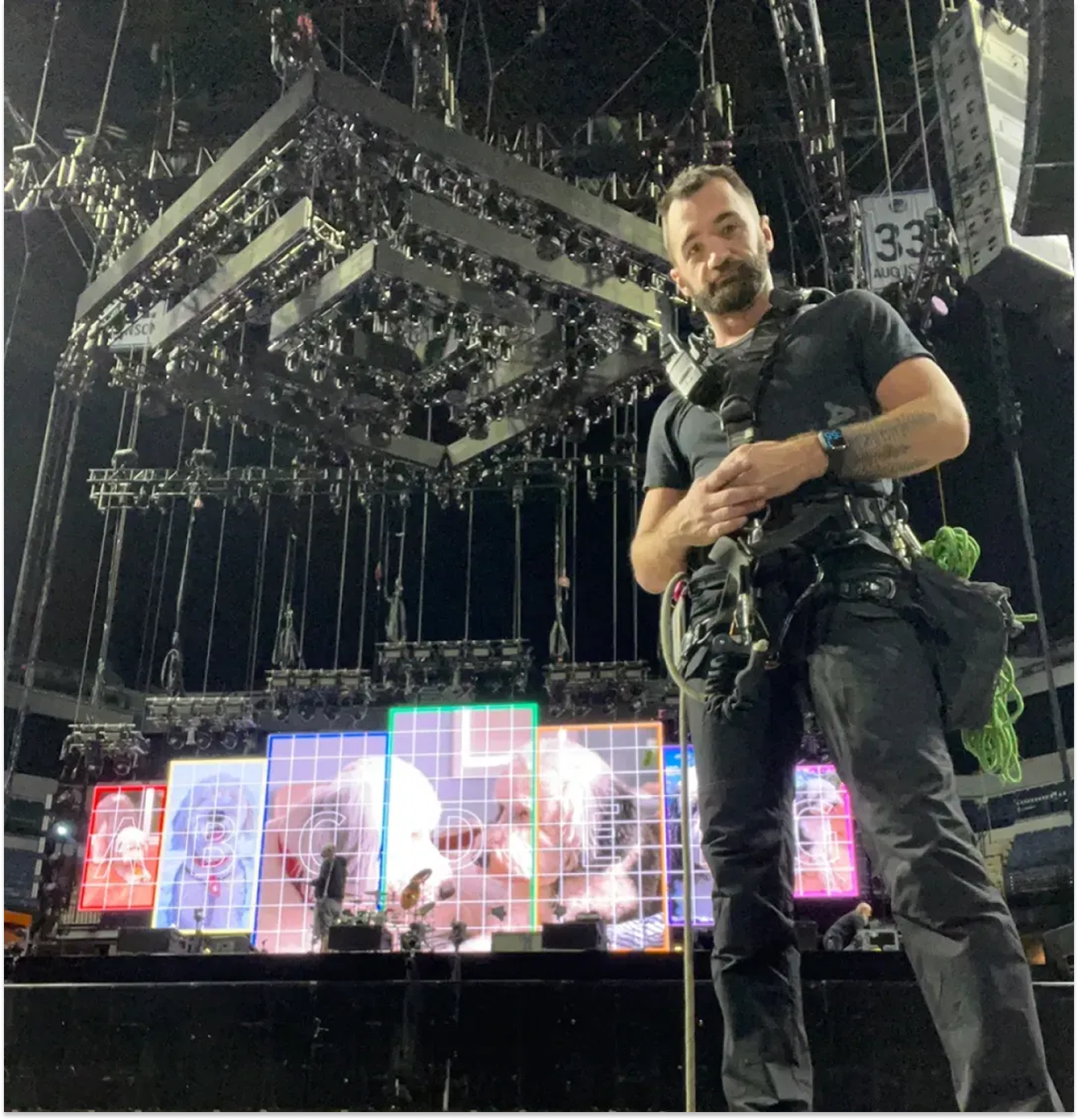 A man with a beard, wearing a black t-shirt and gray pants, stands in front of a large stage with colorful digital screens, holding a camera in an indoor concert or event setting.