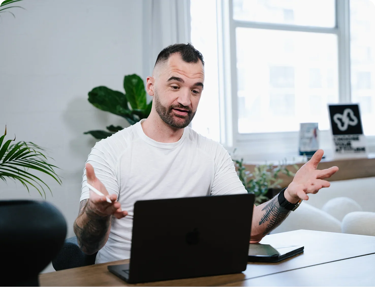 A man with a beard and tattoos on his arms is having a video call or online meeting, gesturing with his hands. He is sitting at a desk with a laptop, a tablet, and a pen, in a bright office space with large windows and some plants in the background.