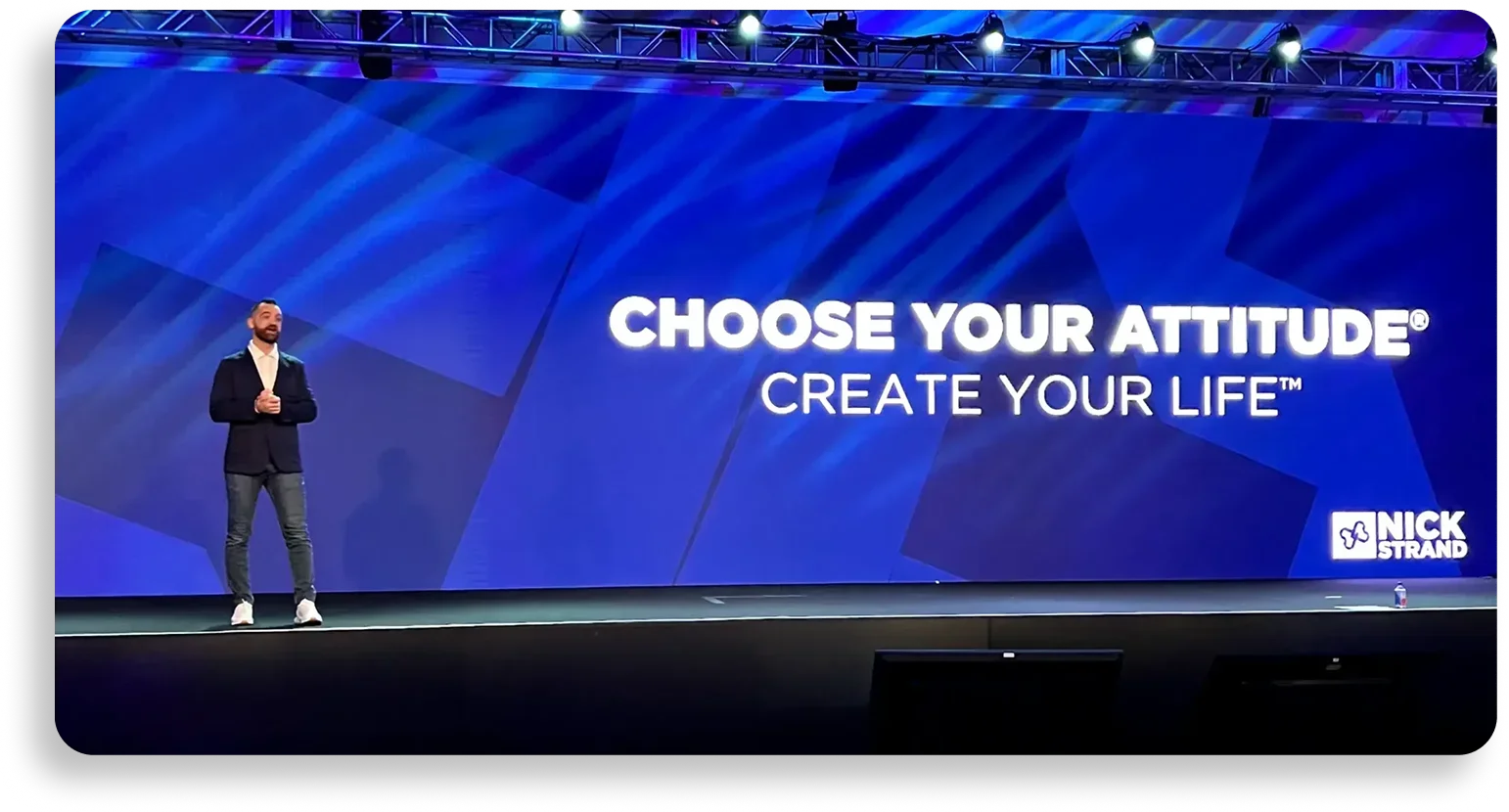 A man stands on a stage with a large blue screen behind him that displays the text 'Choose Your Attitude, Create Your Life' along with a logo for Nick Strand. The man is wearing a navy blazer, light-colored shirt, jeans, and white sneakers. The stage has black flooring, and there are stage lights at the top.