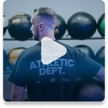 Back of a man at a gym, wearing a shirt that says 'Athletic Dept.', with kettlebells on racks behind him.