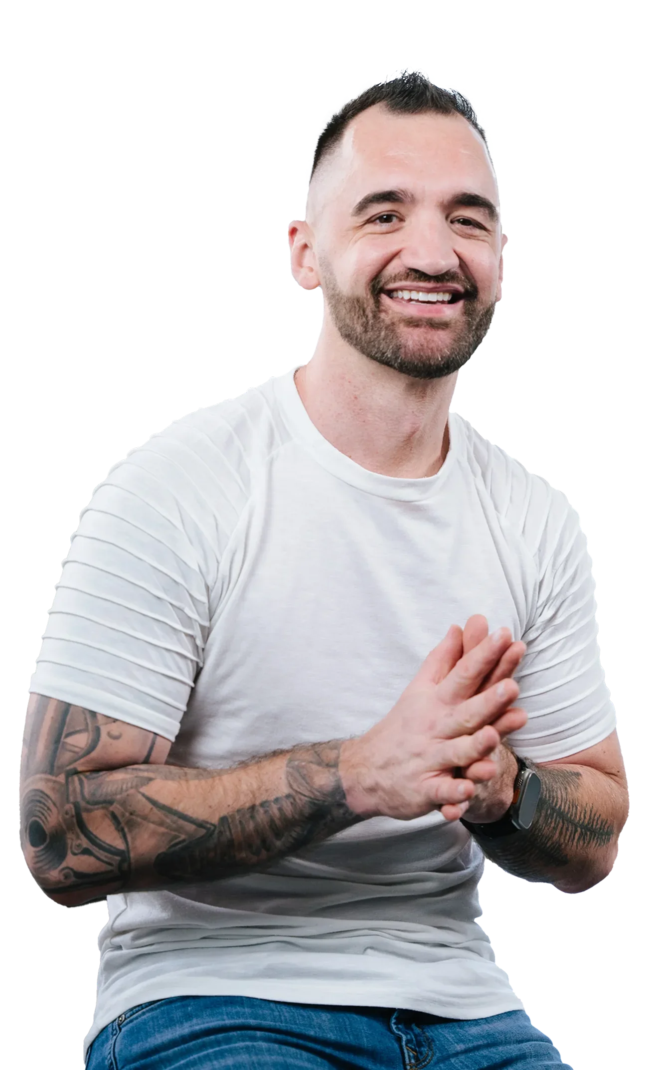 A smiling man with short dark hair, a beard, and tattoos on his arms, wearing a white t-shirt, sitting and holding his hands together in a gesture of contentment or prayer.