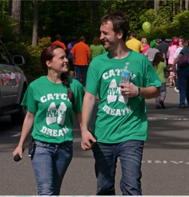 Two people walking together at a charity walk, both wearing green T-shirts that say 'Catch My Breath,' holding hands and smiling, with a group of people in the background.