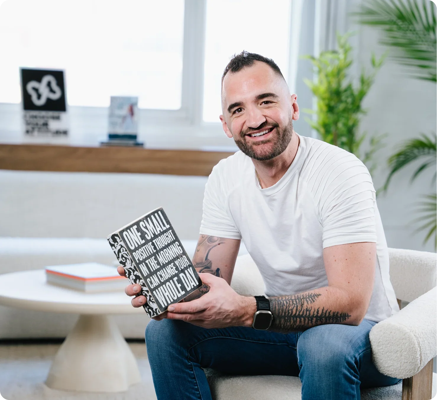 A smiling man with a beard and short dark hair, wearing a white t-shirt, blue jeans, and a smartwatch, sitting on a cream-colored sofa in a bright room with large windows and green plants, holding a book with the cover text 'One small positive thought in the morning can change your whole day'.