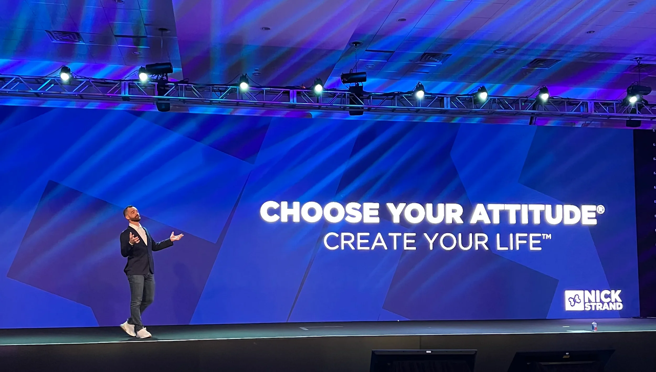 A man in a black blazer, white shirt, and jeans stands on stage with a large blue screen behind him displaying the text 'Choose Your Attitude® Create Your Life™' and the Nick Strand logo. The stage has colorful lighting overhead.