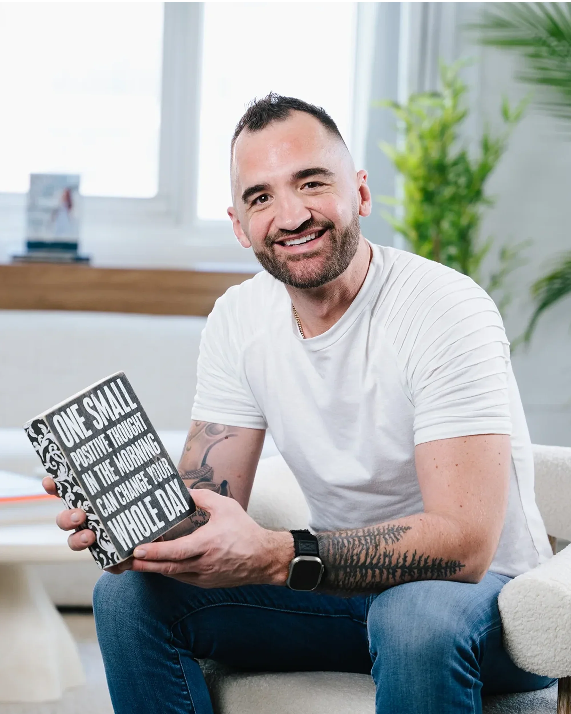 A smiling man with a beard and tattoos on his left arm, wearing a white t-shirt, sitting on a beige sofa in a bright room, holding a black and white motivational quote mug that reads, "One small positive thought in the morning can change your whole day."
