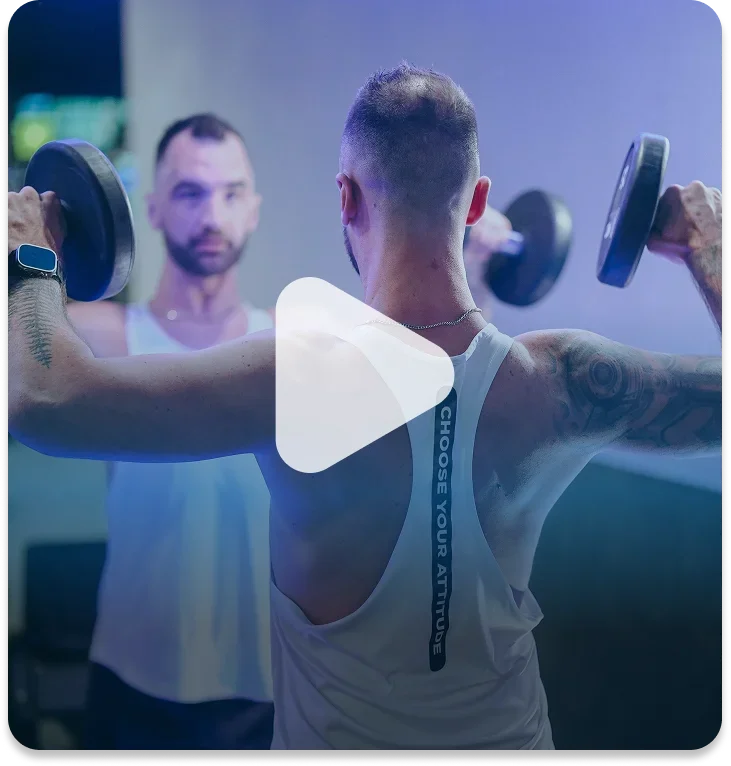 Man using dumbbells for shoulder exercise with trainer observing in the background.