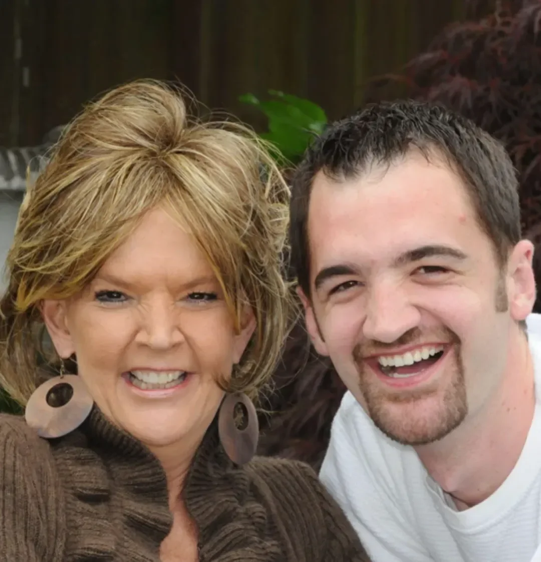 A smiling woman with blonde hair and large earrings taking a selfie with a smiling man with short dark hair. They are outdoors with plants in the background.