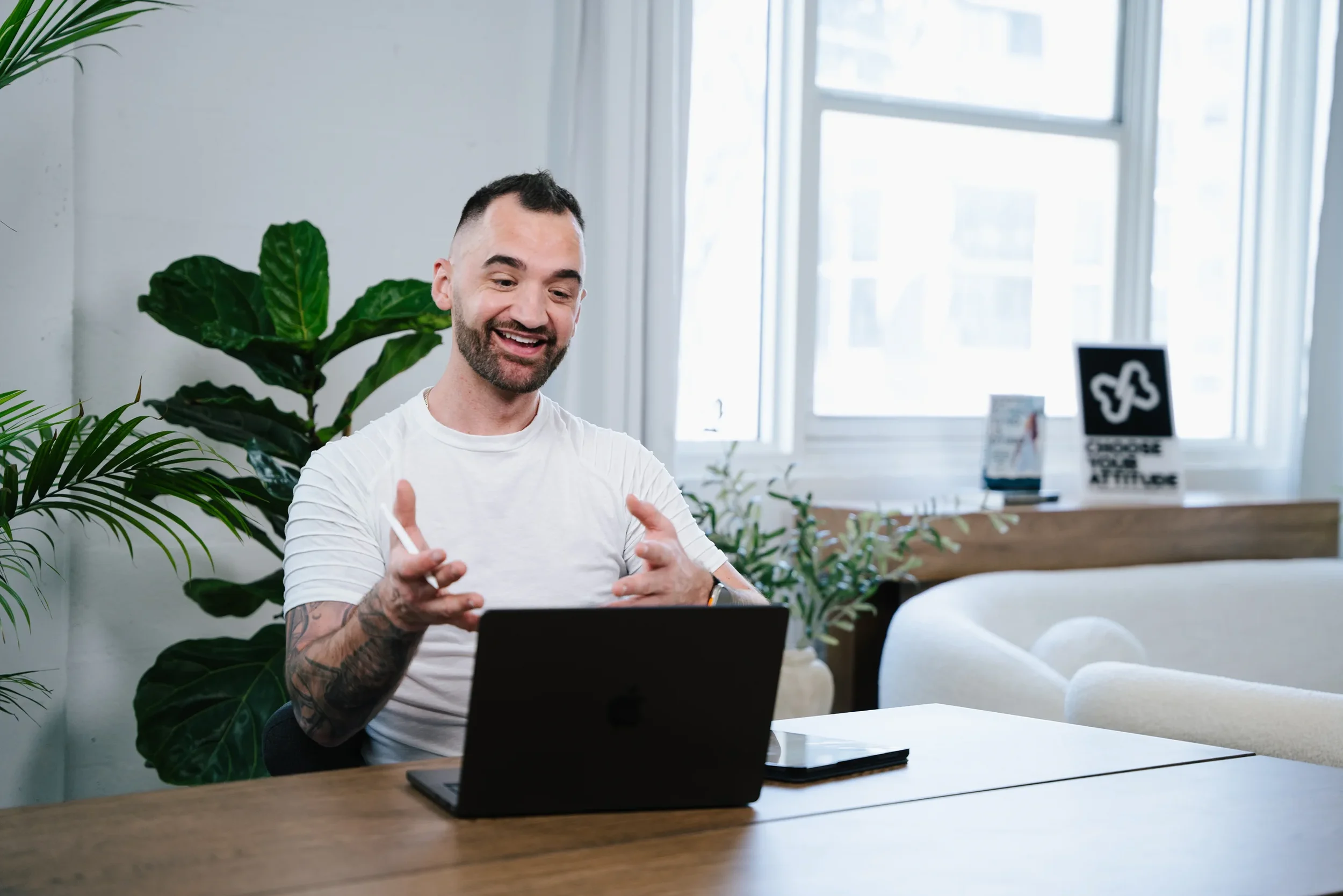 Man with a beard wearing a white T-shirt sitting at a wooden table in a bright, modern office, smiling and gesturing with his hands while looking at a laptop.
