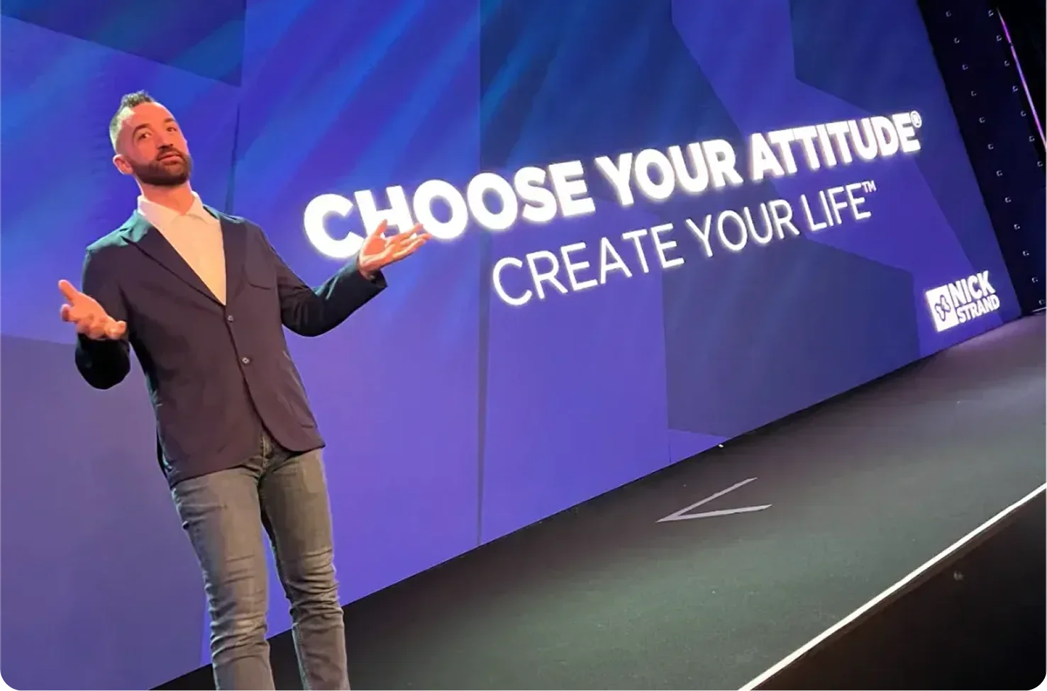 Man in blazer gesturing on stage in front of a large screen displaying the text 'Choose Your Attitude, Create Your Life' and the Nick Strand logo.