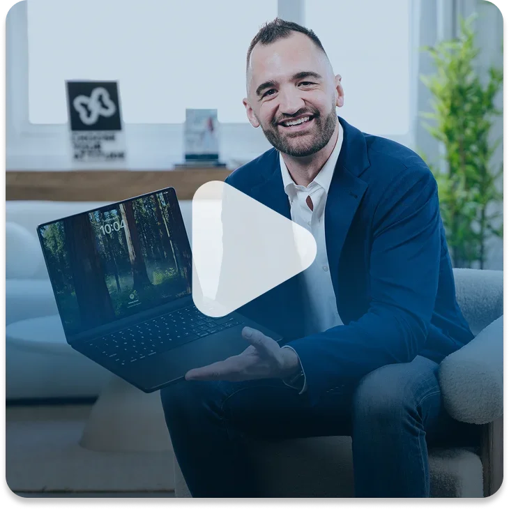 A man in a blue suit sitting on a gray couch, holding a laptop showing a forest scene on the screen, smiling at the camera in a modern office setting.