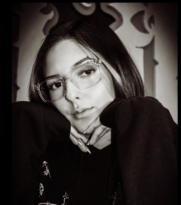 A young woman with short dark hair and clear glasses resting her chin on her hands, looking at the camera, in a black hoodie, with decorative wall patterns in the background.