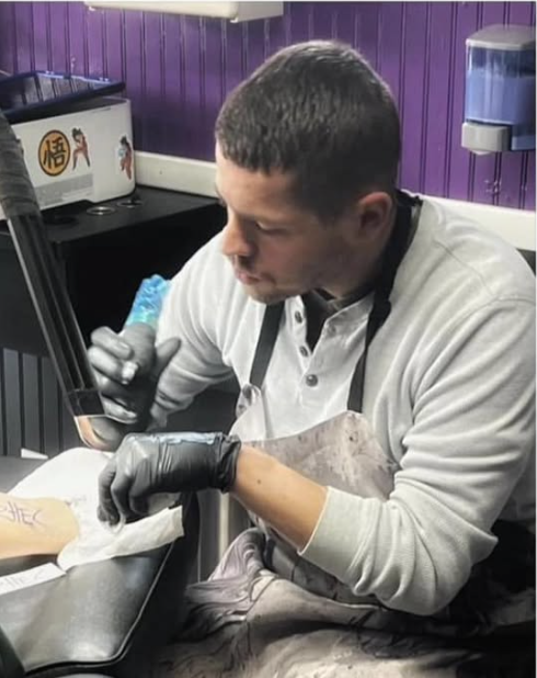 A man wearing a white shirt, black gloves, and an apron is sitting at a table, looking at his phone, in a purple-walled room with a water dispenser in the background.
