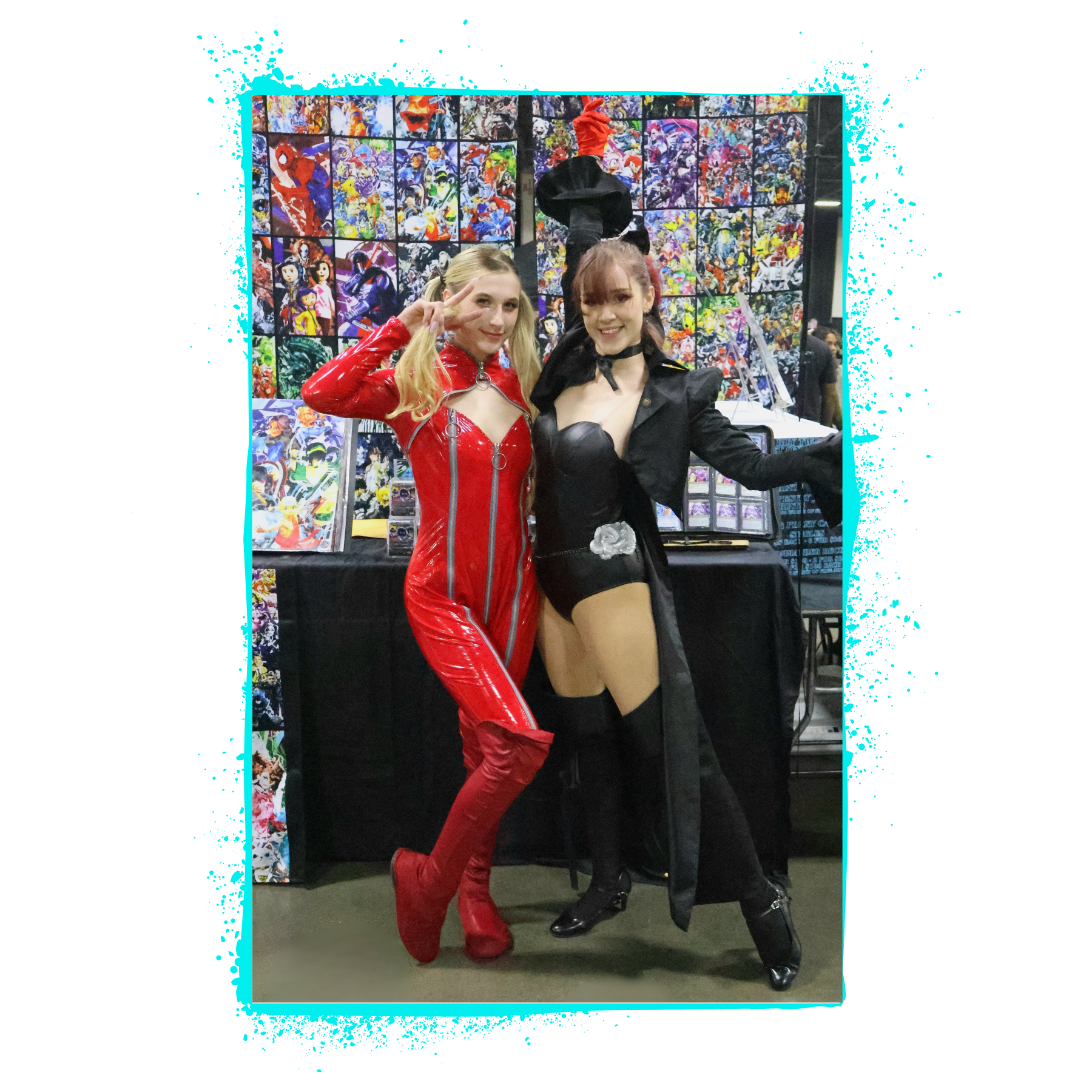 Two women in cosplay costumes posing at a comic convention. One dressed as a red latex character with blonde hair, and the other in a black outfit with a hat and dark hair. They are standing in front of a table displaying artwork.