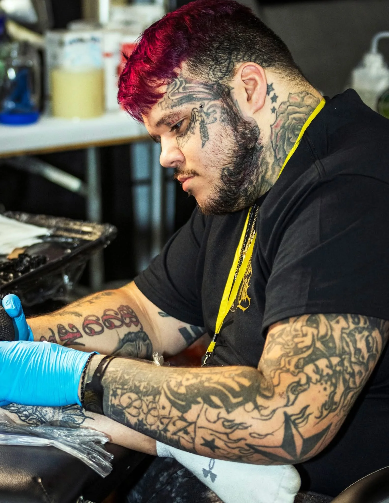 A man with red and black hair and numerous tattoos is getting a tattoo on his arm in a tattoo parlor.