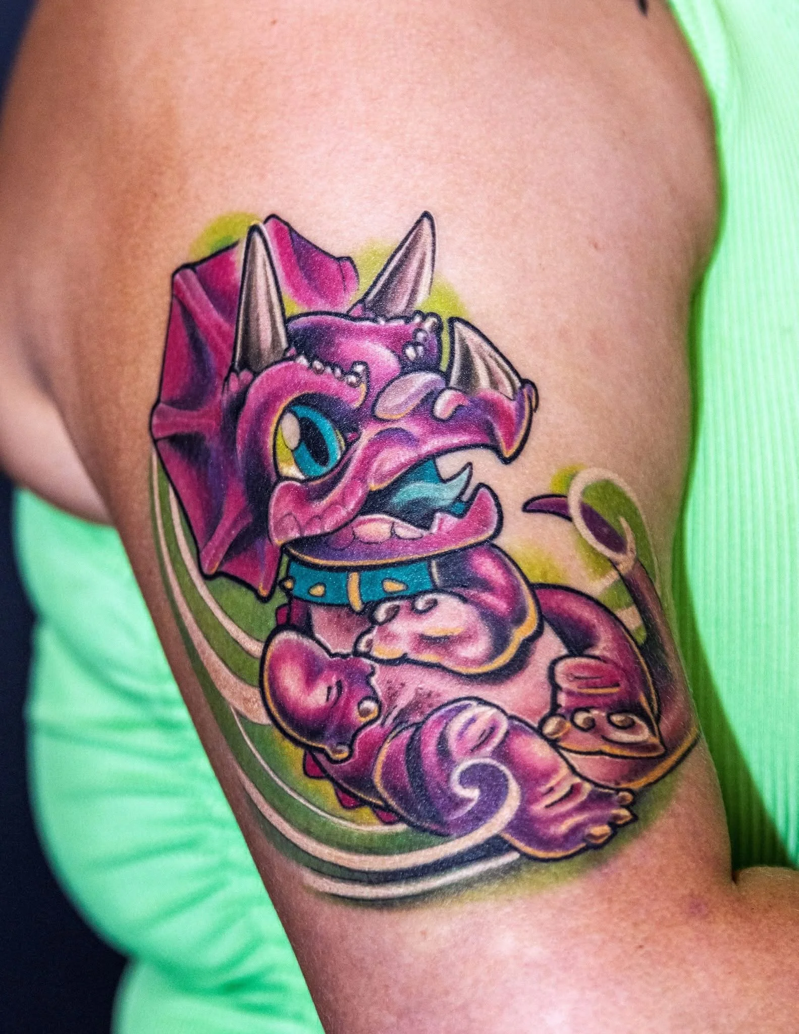 Colorful tattoo of a stylized pink and purple dragon puppy with large blue eyes, wearing a blue collar, on a person's upper arm.
