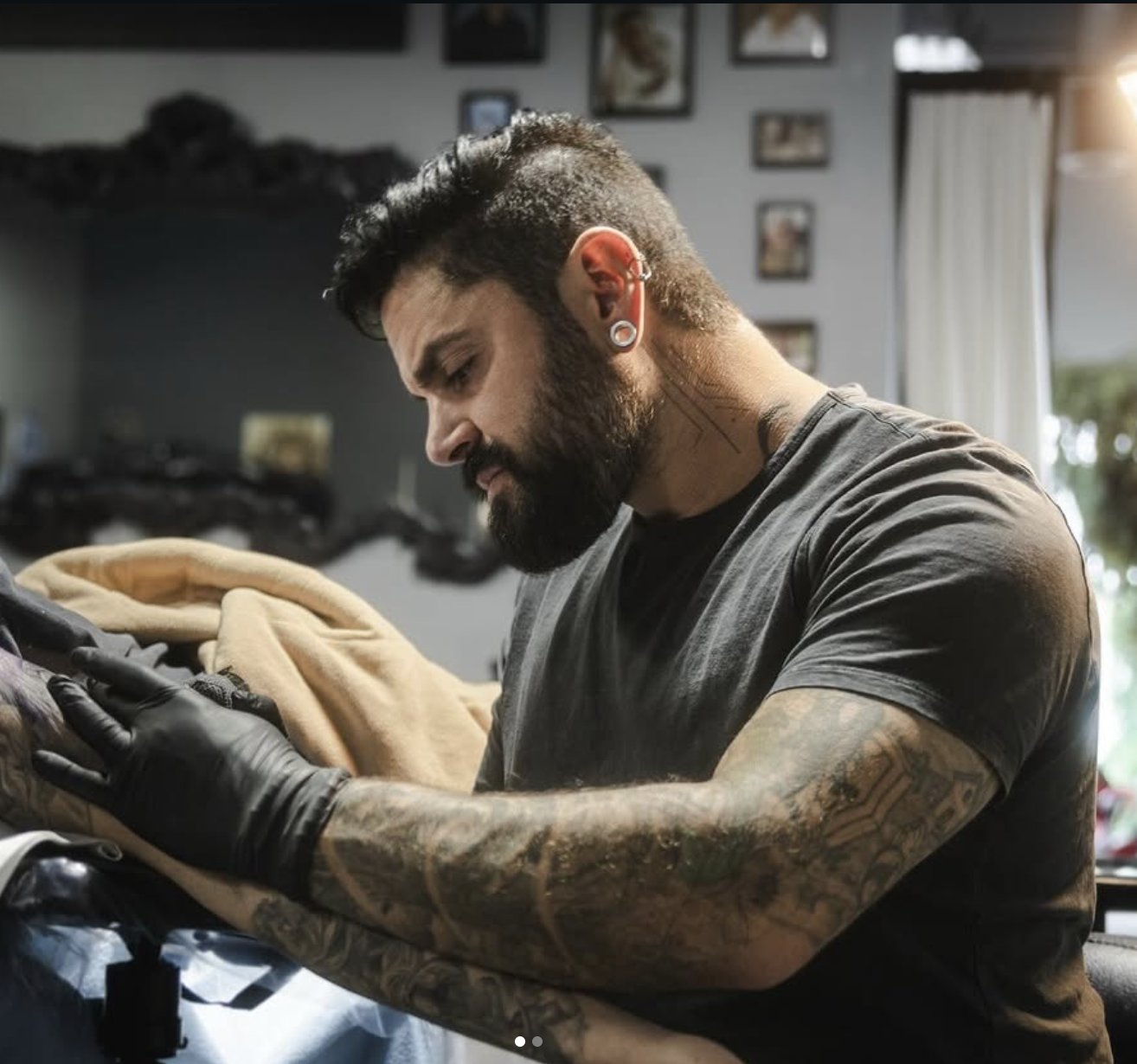 Tattooed man with earrings and black gloves tattooing a client's arm in a tattoo parlor.