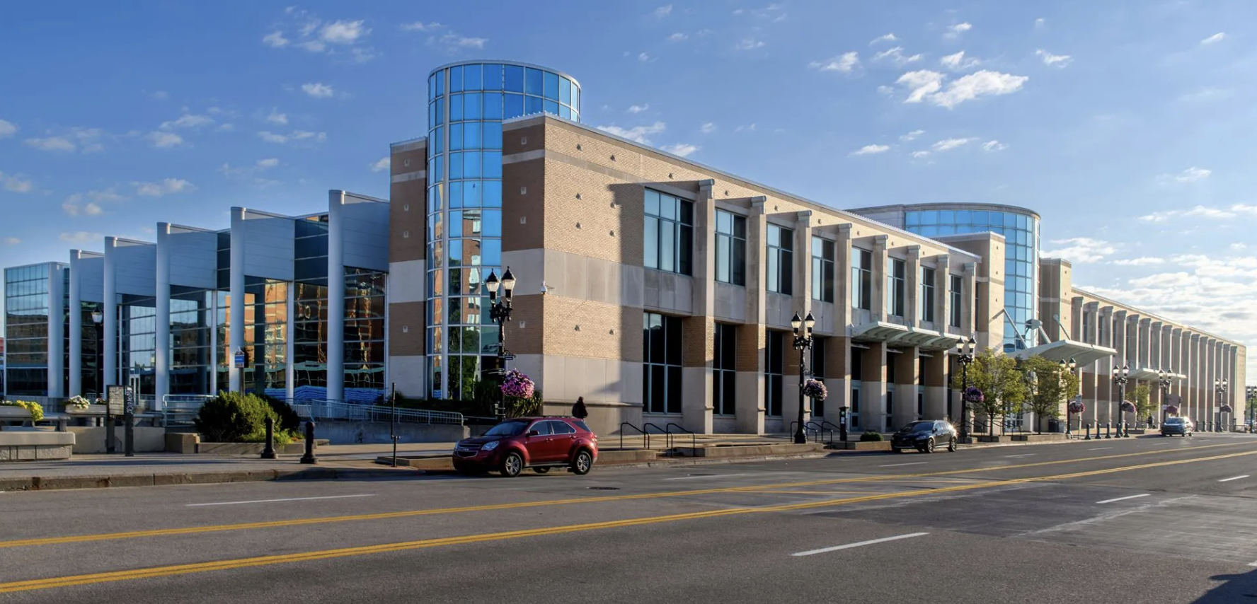 Modern building with glass windows and brick facade, street with cars parked and pedestrians, flowering plants, lamp posts, under a partly cloudy sky.
