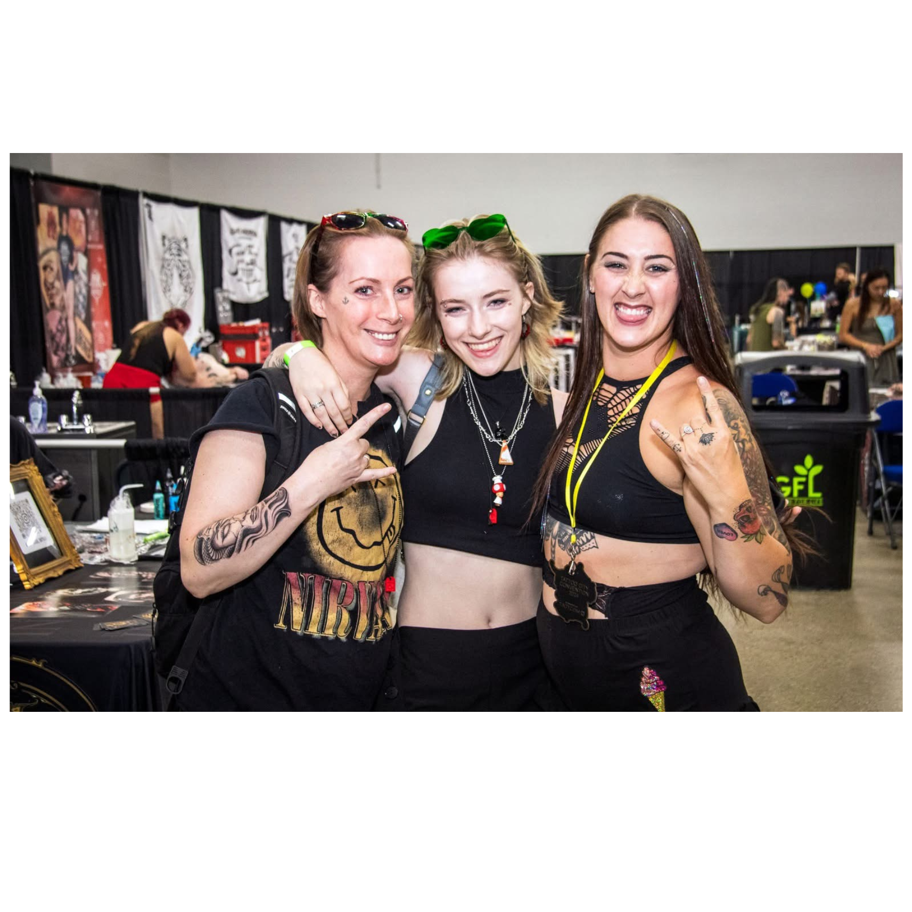 Three women smiling at a convention, with booths and artwork in the background, showing casual and rebellious style.