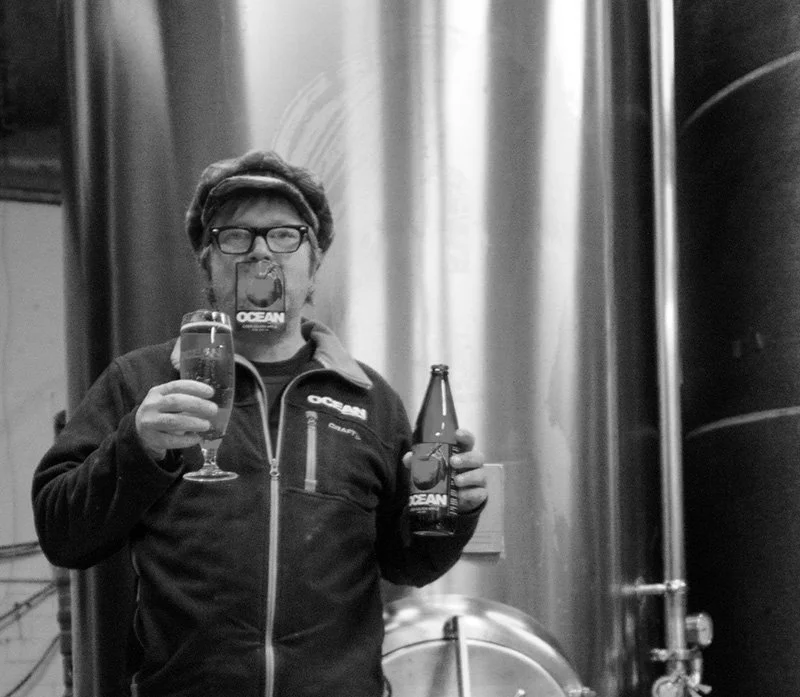 Person som håller ett öl-glas och en öl-flaska i en bryggerimiljö, fotograferad i svartvitt.