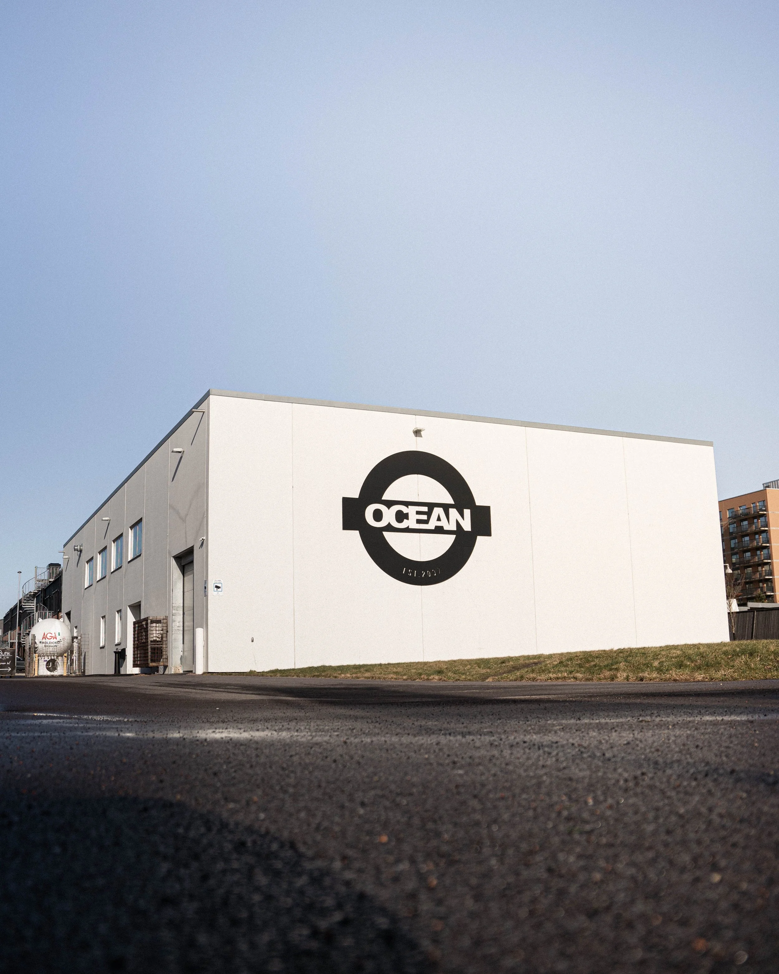 En vit industribyggnad med en stor svart och vit logo som säger 'OCEAN' och 'EST. 2017' på ena väggen, mot en klar blå himmel.