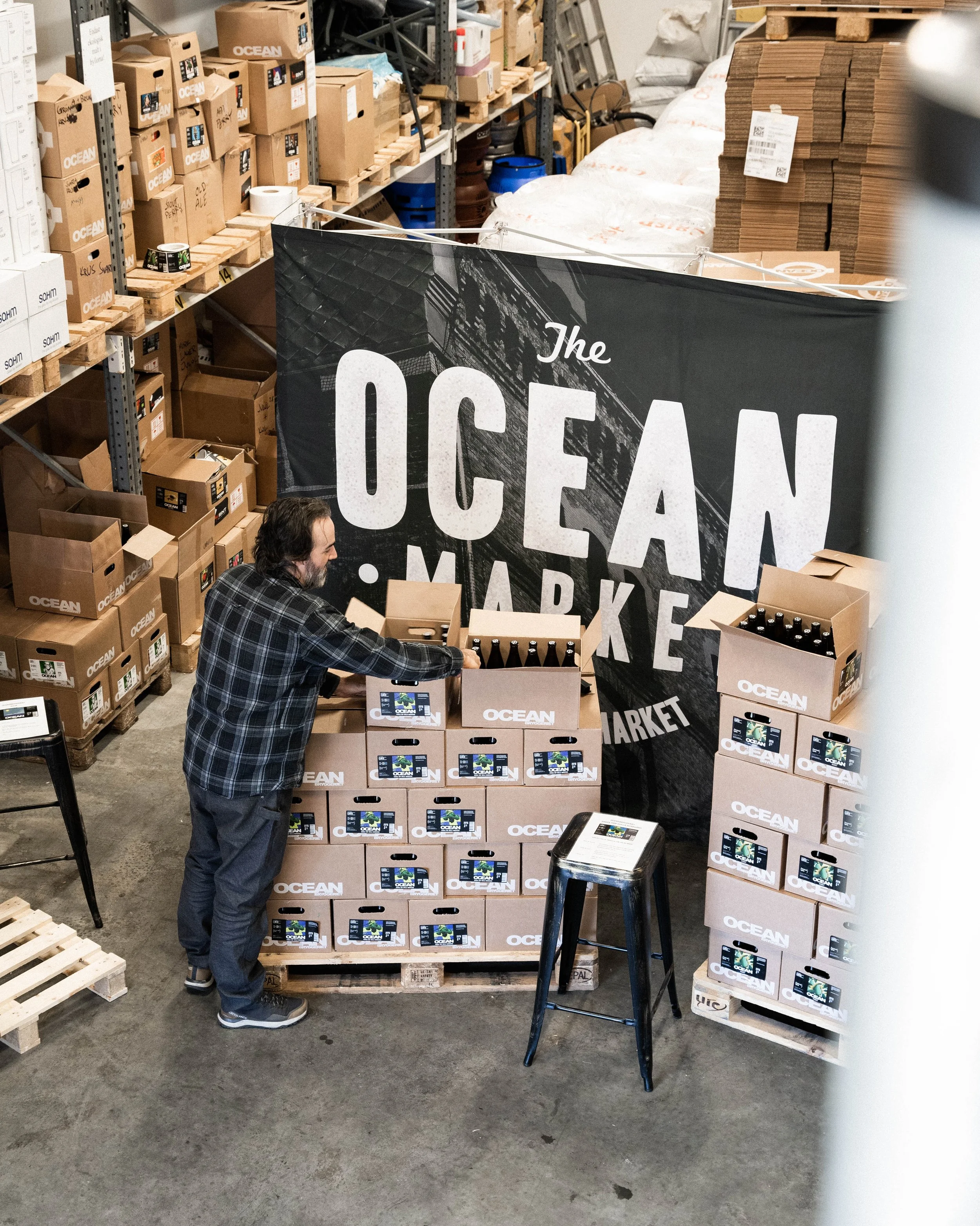 En man i en lagerlokal packar ölflaskor i kartonger med Ocean-märke framför en stor skylt som säger 'Ocean Market'. Lagerhyllor med tomma och fyllda kartonger finns i bakgrunden.