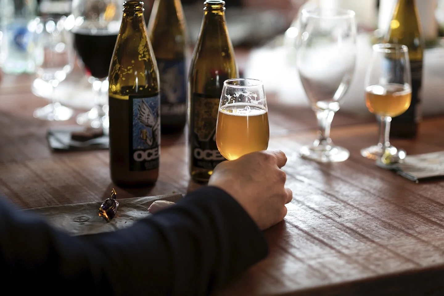 En hand som håller ett glas öl på ett träbord med flera flaskor och glas med öl och vin i bakgrunden på gårdsförsäljning, restaurang.
