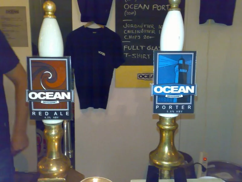 Två ölkrus med etiketter för Ocean Bryggeri, ett svart T-shirt och en svart tröja hängande på väggen i bakgrunden, samt en svart tavla med vit text.