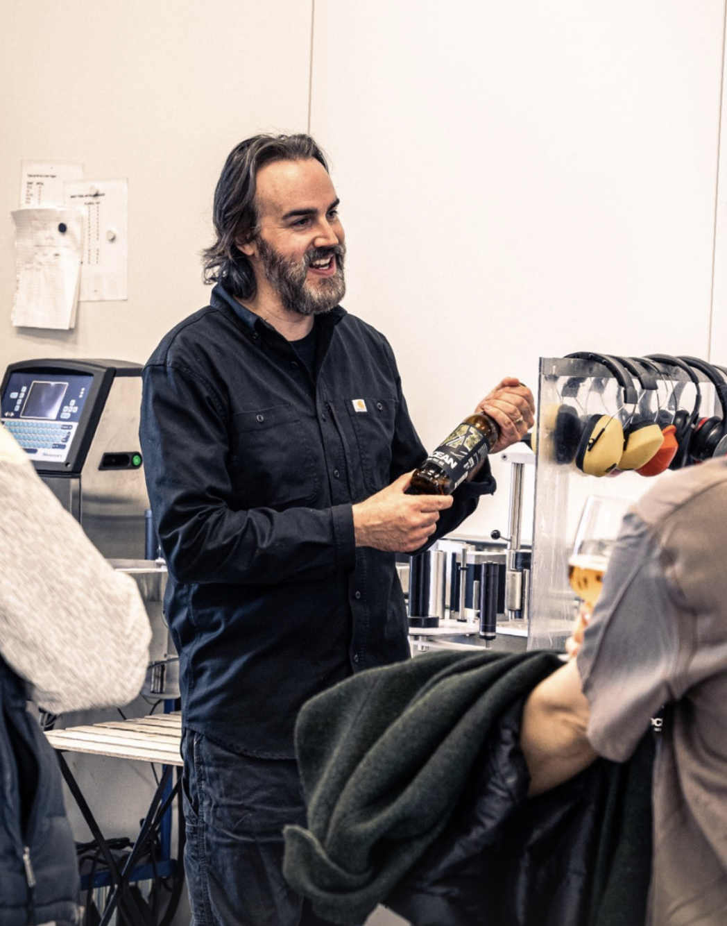 En ölbryggare som håller en ölflaska och talar inför en publik i ett bryggeri