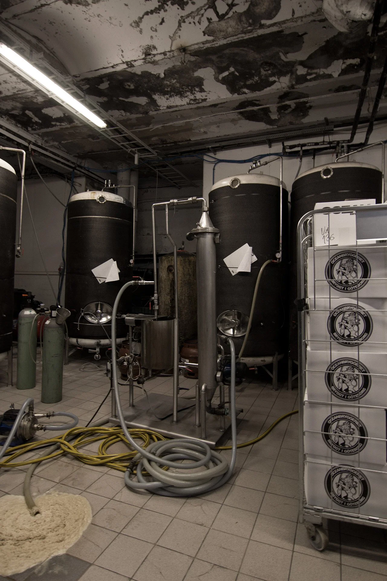 En bryggerilokal med tre stora svarta tankar, kopparkärl och slangar. Det finns även gasflaskor och ett rullvagn med öl- eller bryggerimärke.