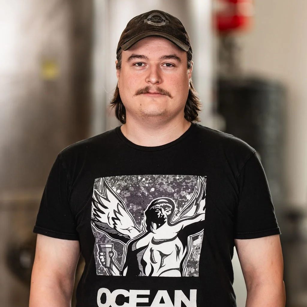 Man med hatt och skäggstubb, svart T-shirt med grafisk bild av Poseidon med vingar och en öl, står i en industrimiljö.