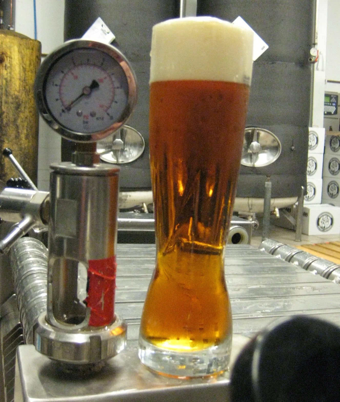 Ett glas öl med skum på toppen, bromsmätare och andra bryggeriutrustning i bakgrunden.