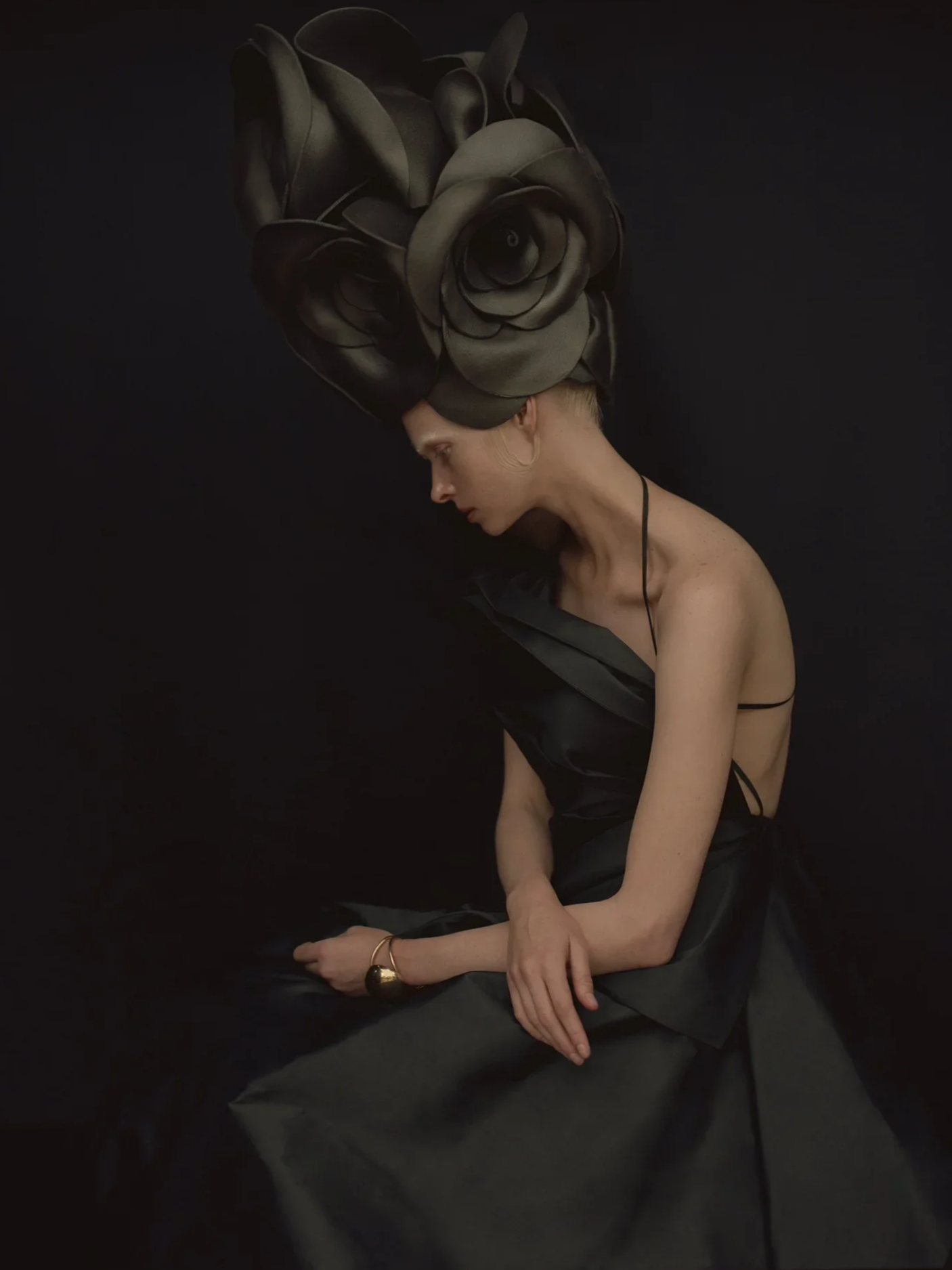 Aliona  with a large, sculptural black floral headpiece, wearing a black satin dress, seated against a dark background, looking downward, for Reserved Magazine.