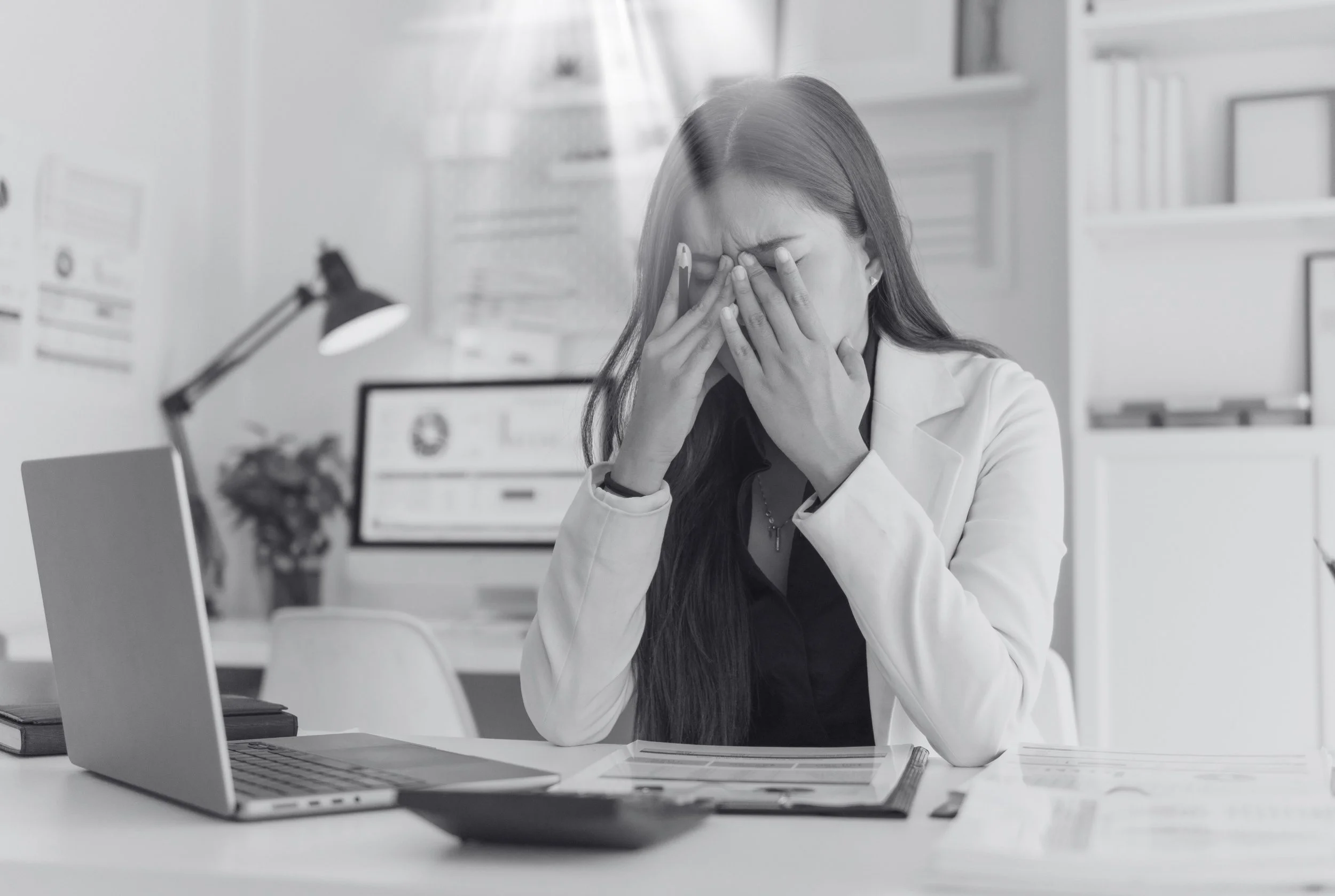 Une femme d'affaires a l'air stressée ou fatiguée, assise à un bureau avec un ordinateur portable, des documents et une lampe de bureau dans un environnement de bureau.