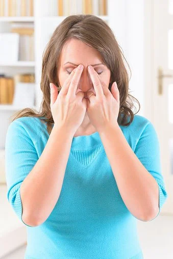 Femme anxieuse se tenant le visage, yeux fermés dans un intérieur lumineux.