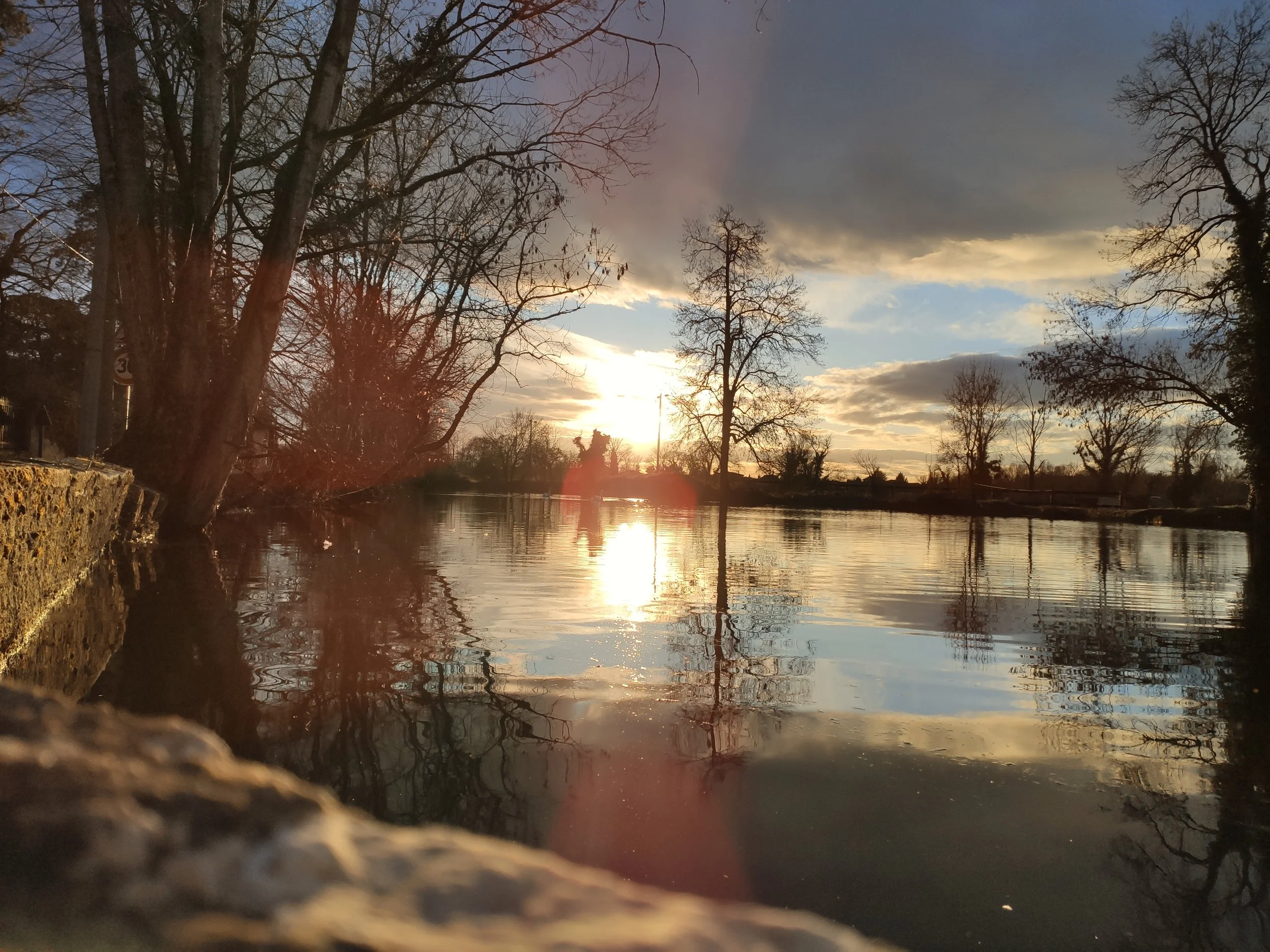 Coucher de soleil sur une rivière bordée d'arbres nus, reflet brillant du soleil sur l'eau, ciel partiellement nuageux.