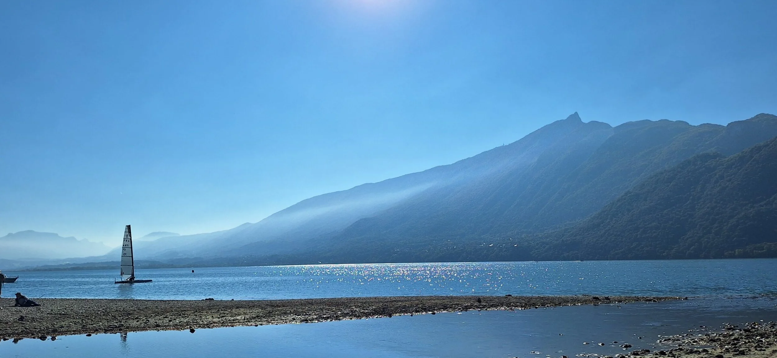 Lac avec montagnes en arrière-plan, un voilier et un petit bateau en premier plan, ciel clair en journée.