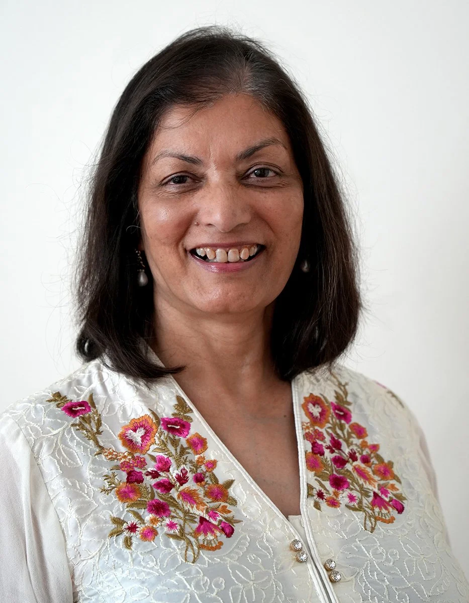 A woman with shoulder-length dark hair, smiling, wearing a white embroidered blouse with pink and orange floral designs and pearl earrings.