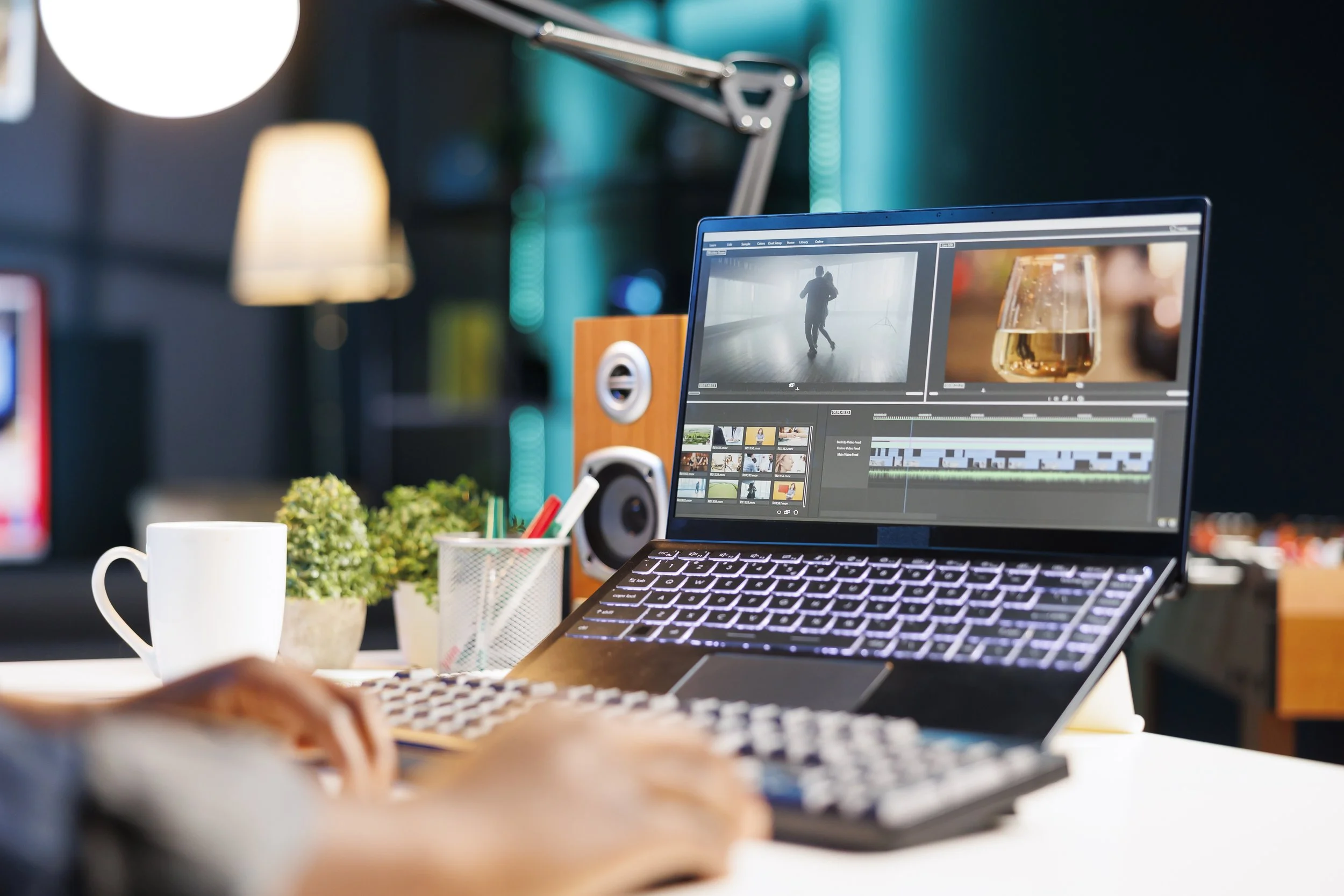 A person working on video editing on a laptop in a modern workspace, with a desktop monitor displaying editing software, a coffee mug, and small potted plants on the desk.