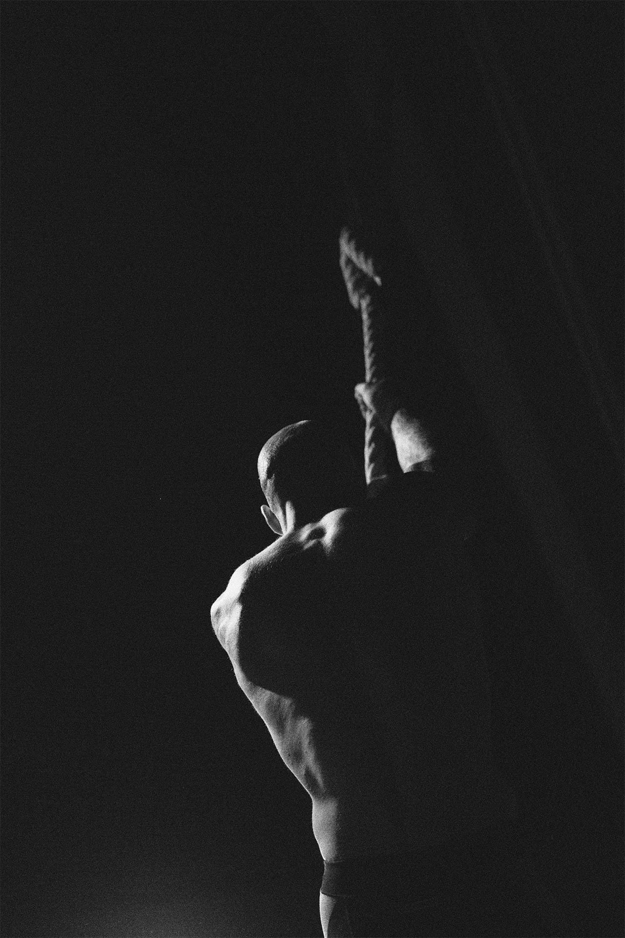 Hombre musculoso en postura de musculación o ejercicio, visto desde atrás en un ambiente oscuro, en blanco y negro.